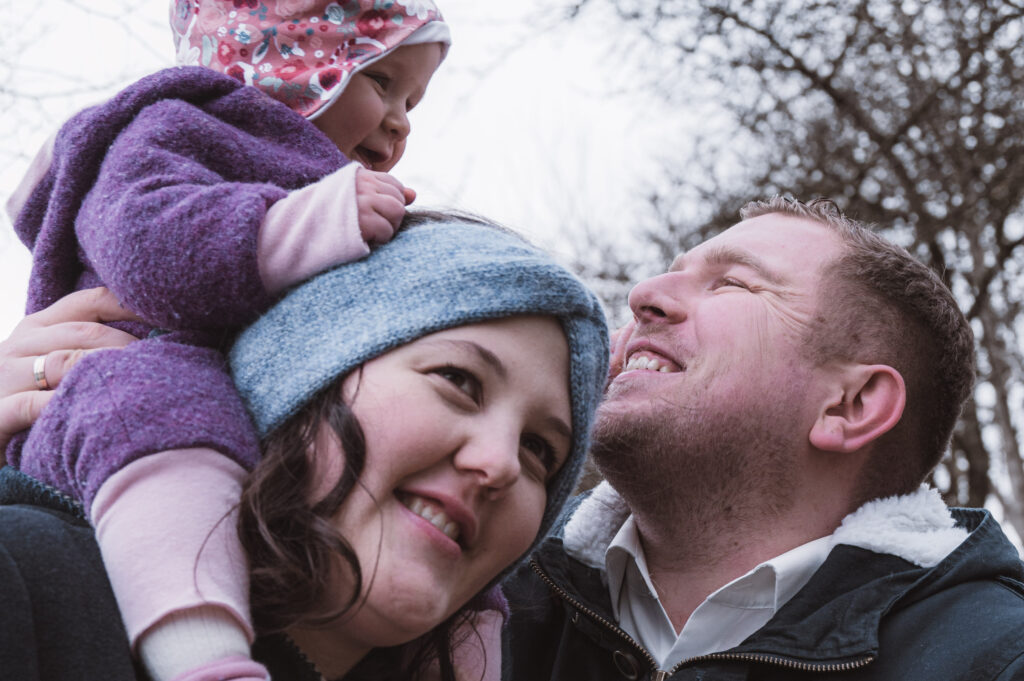 Familienshooting mit Mama, Papa und kleinen Kind by Carografie Caroline Nagl im schönen Wieselburg. Der Vater lächelt das Kind an, welches auf der Mama sitzt